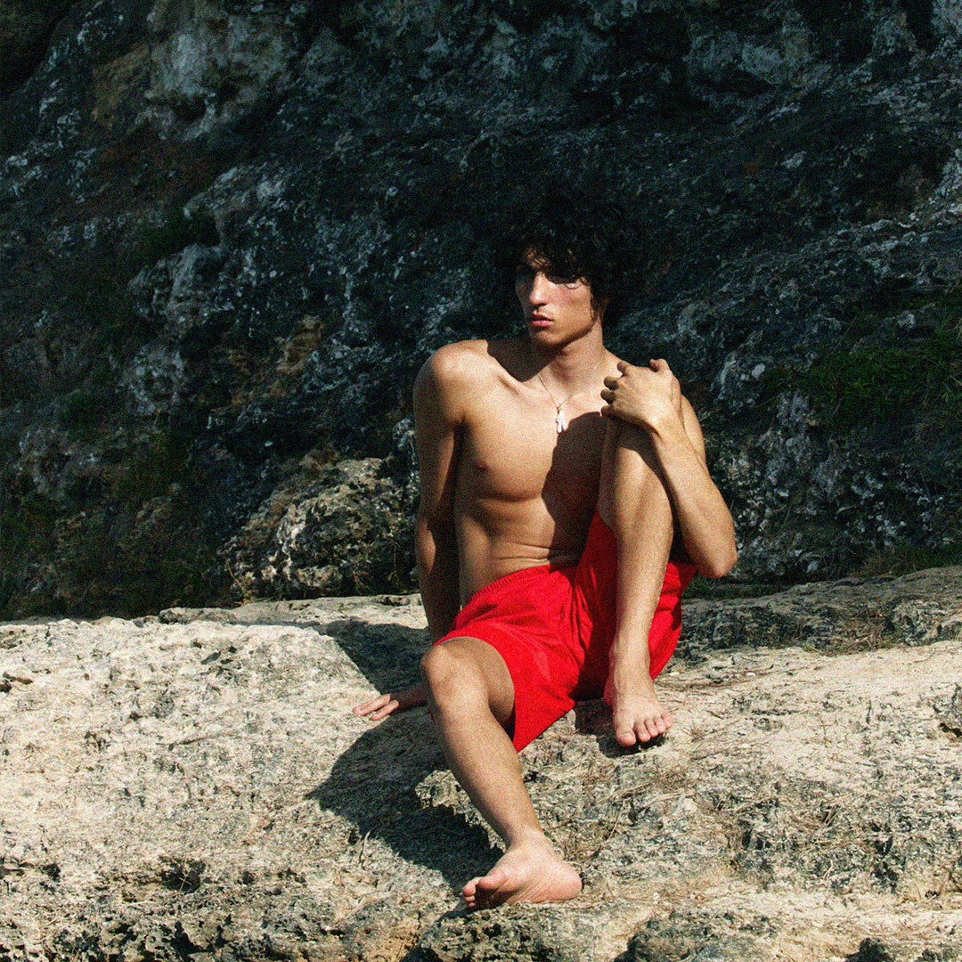 Boy in red swimming trunks sitting uncomfortably on a large rock.